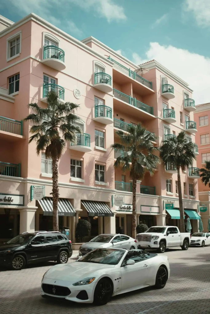 A vibrant street scene in downtown Boca Raton featuring a pastel pink building with green balconies, palm trees, and a variety of parked cars, including a white convertible sports car in the foreground.