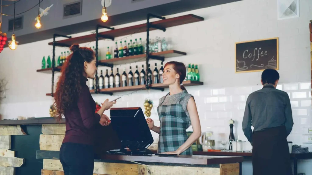 A cozy coffee shop interior with a customer interacting with a barista at the counter, surrounded by shelves of bottles and warm lighting.