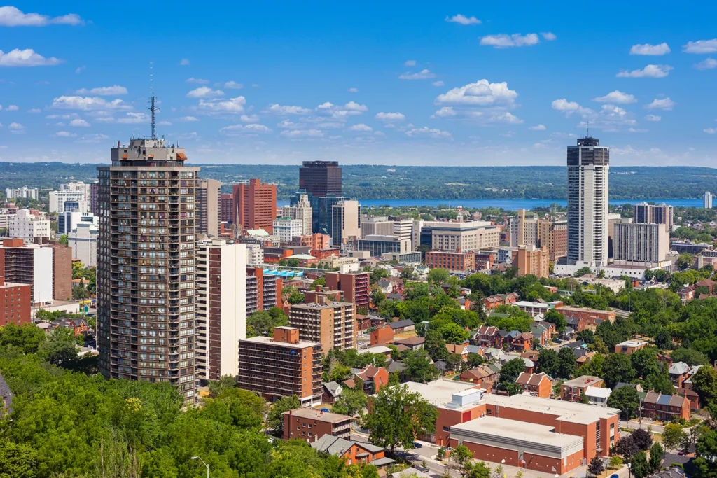 An aerial view of Hamilton's skyline with high-rise buildings, lush green trees, and Lake Ontario in the background under a clear blue sky taken by best local seo agency in Hamilton.