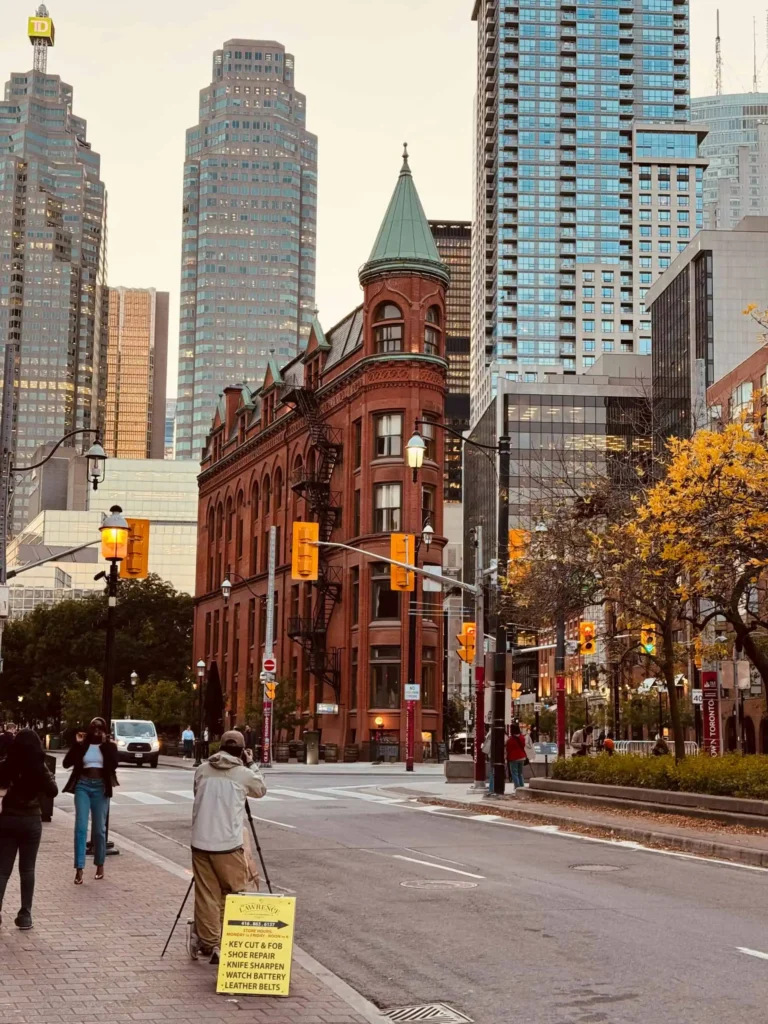 A bustling Toronto street scene featuring a historic red-brick building, modern skyscrapers, and a person with a sign offering services, with autumn trees in the background.